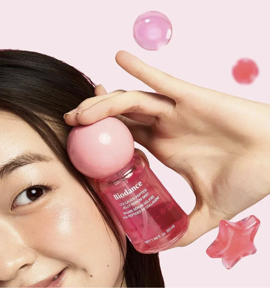 Model houdt de roze flacon Biodance Collagen Peptide Jelly Serum Mist naast haar gezicht, tegen een lichtroze achtergrond.