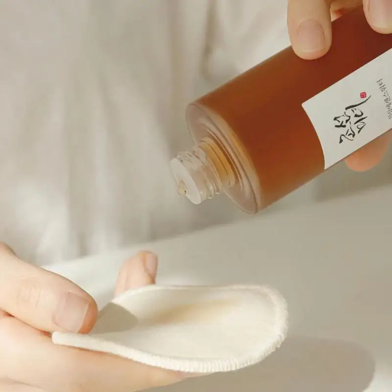 Beauty of Joseon Ginseng Essence Water Essencewater wordt op een witte pad gegoten; bruine fles en handen